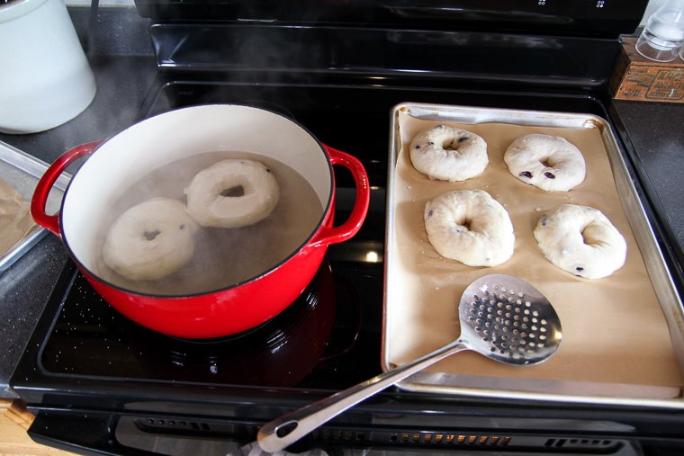 two bagels boiling in a red pot of water sitting next to a baking sheet with the bagels that have already been boiled