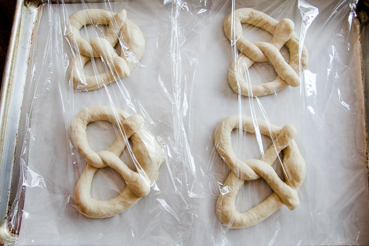 4 unbaked soft pretzels on a baking sheet covered in plastic wrap to proof
