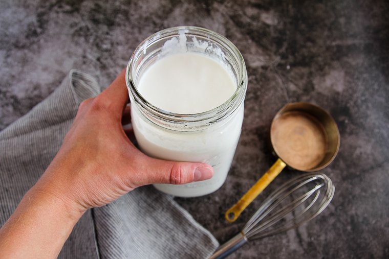 a hand holding a jar of homemade buttermilk next to an empty measuring cup and a whisk