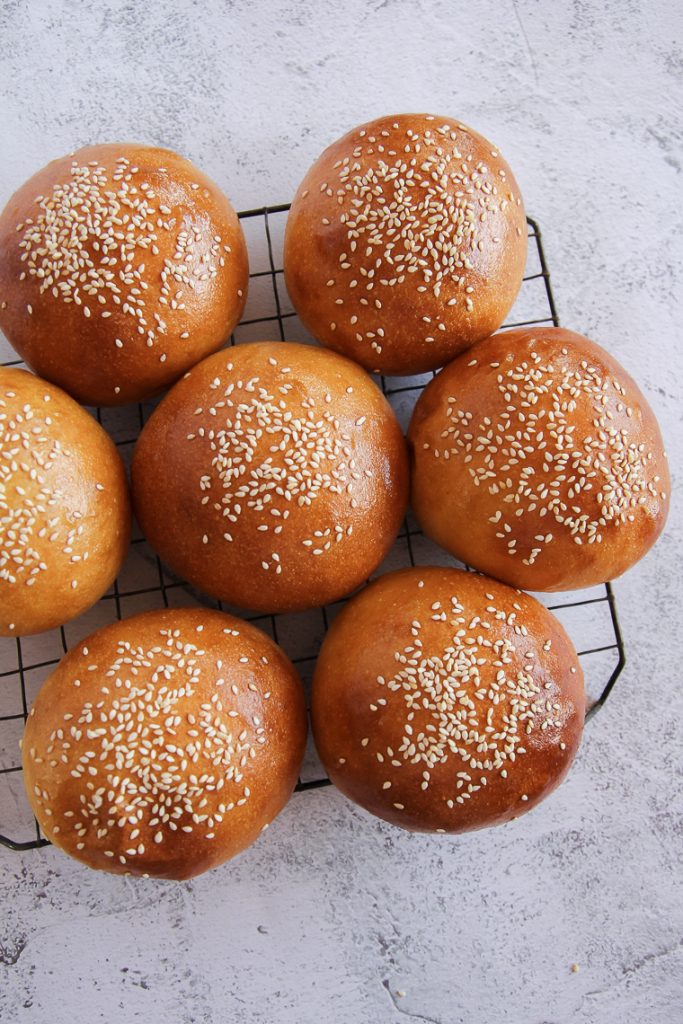 seven sourdough hamburger buns on a cooling rack with a gray background